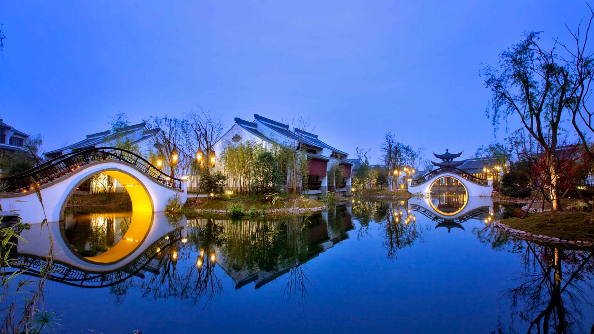 Twilight scene of traditional buildings with curved moon-gate bridges reflected in still blue water and a glowing lantern, calm navy and gold tones