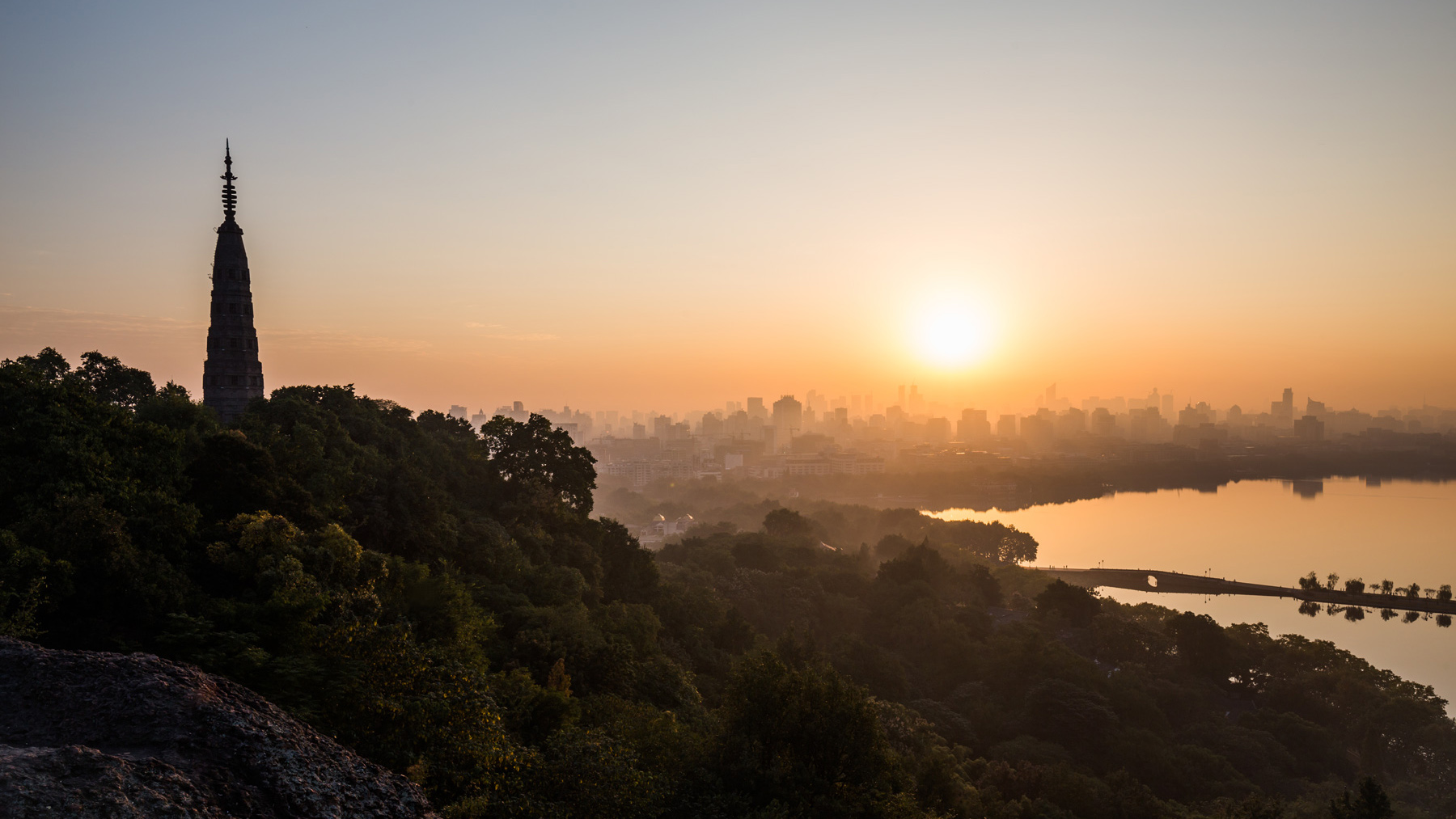 Silhouette of a tall pagoda on a tree-covered hillside overlooking a lake and hazy city skyline at sunrise, warm orange
