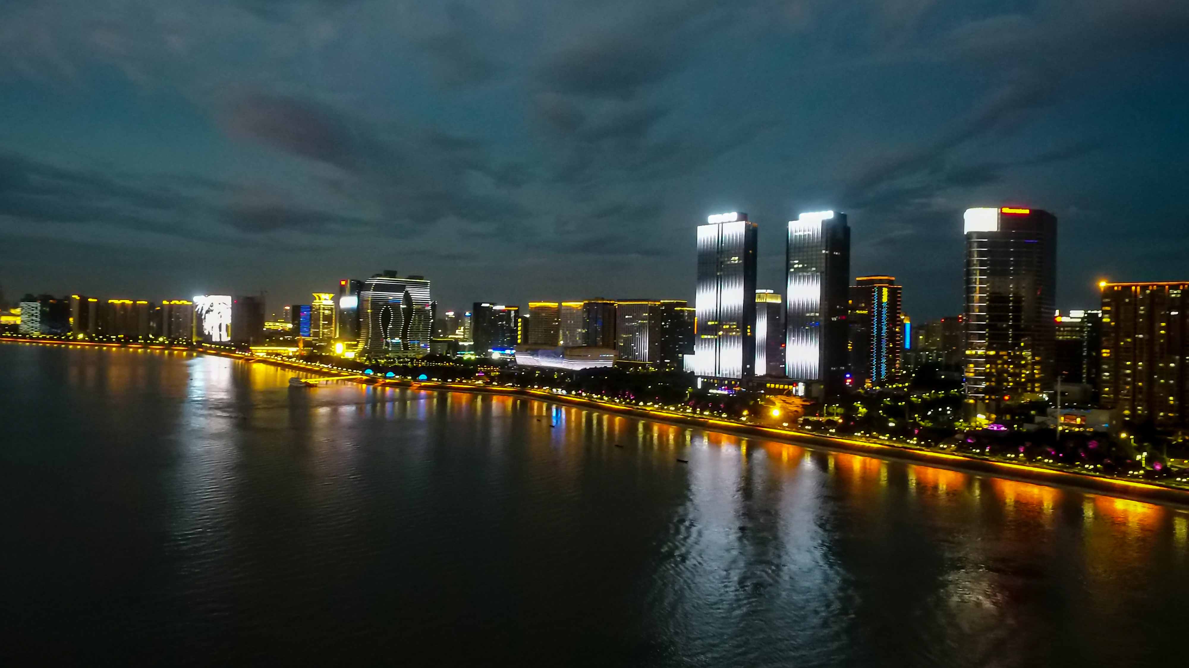 Night cityscape with brightly lit high-rise towers along a waterfront, reflections shimmering in dark water under a cloudy night sky