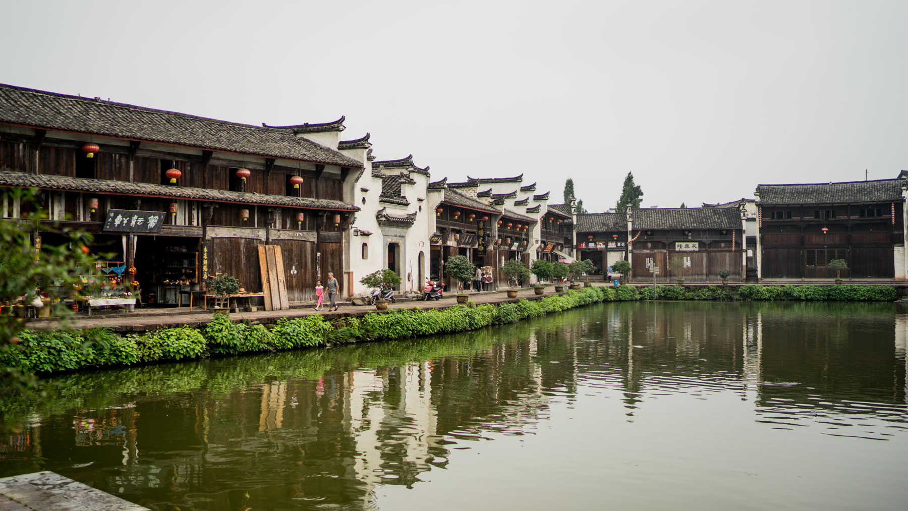 Row of traditional two-story buildings with lanterns along a calm river, green foliage lining the water, peaceful daytime mood