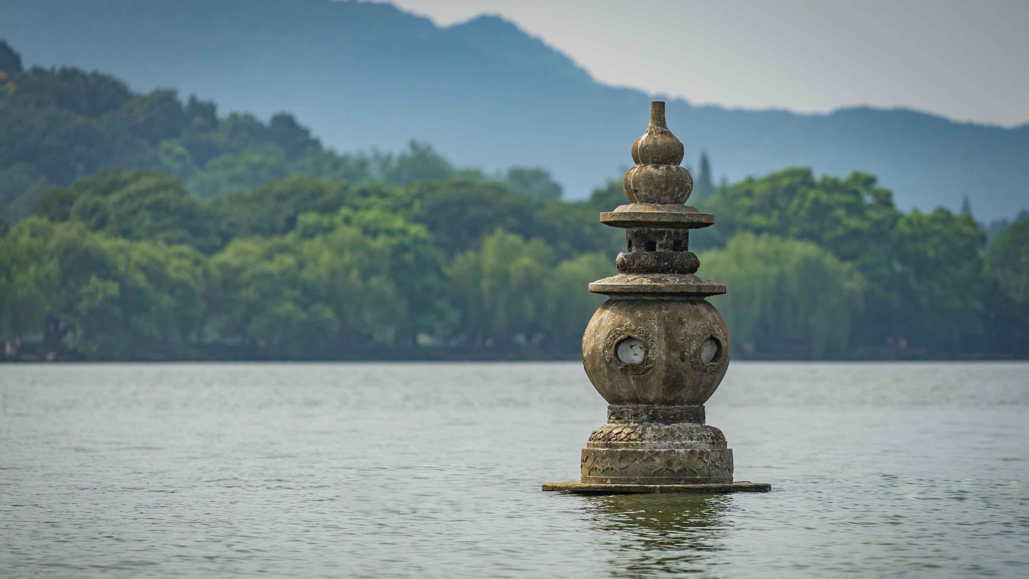 Stone pagoda-shaped monument standing in calm lake water with forested hills in the background