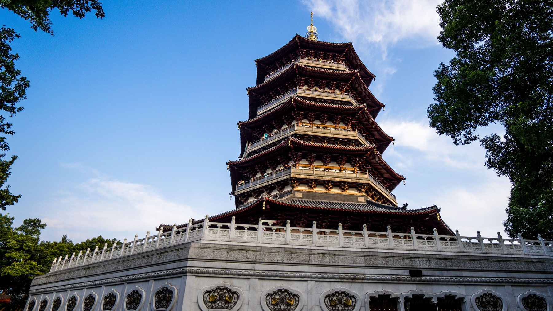 A tall multi-tiered pagoda with dark wooden roofs and gold trim, seen from below against a blue sky with tree branches framing the view